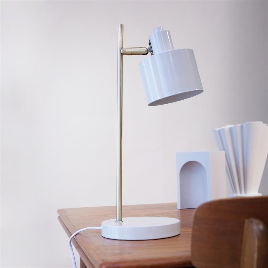 Table lamp stands on a wooden table; the lampshade is tilted forward, mounted on a slender, gold-colored rod with a round white base; neutral wall and decorative white objects behind.