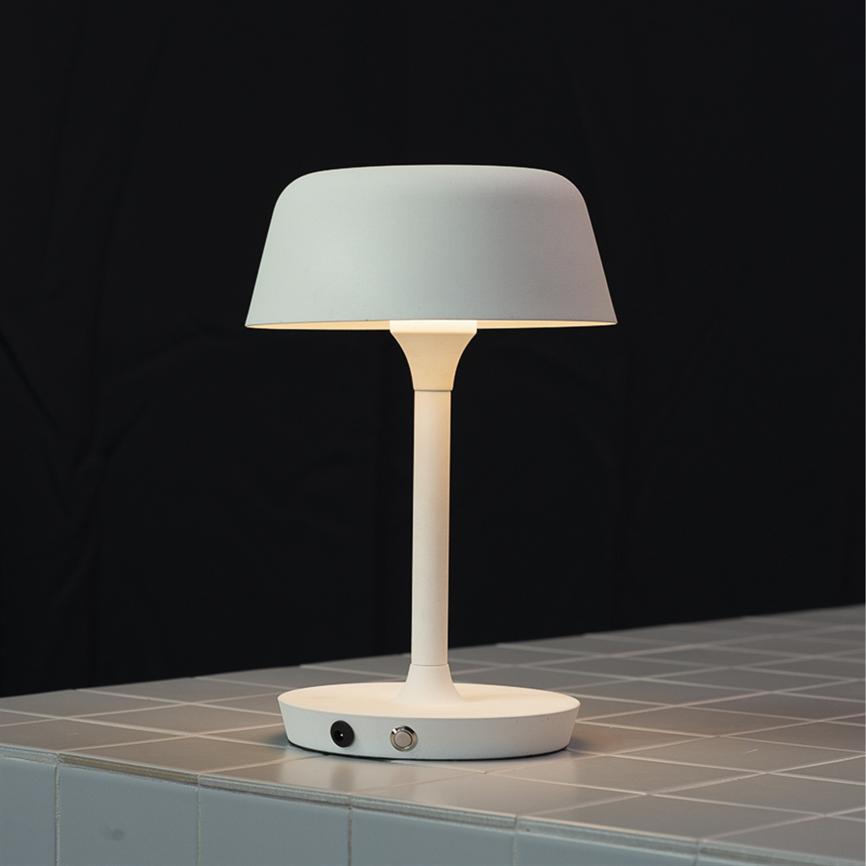 A white, modern table lamp with a round shade stands and shines dimly downwards on a light grey, tiled tabletop in front of a black background.