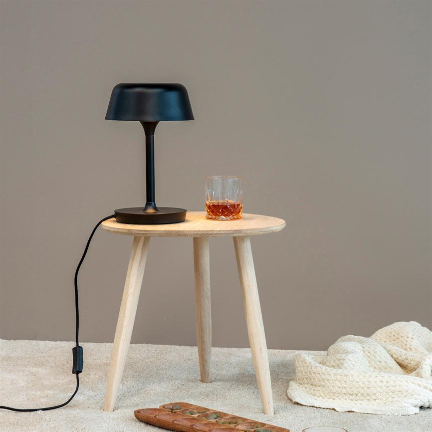 A black table lamp stands on a round light wooden table next to a glass of dark liquid; the cable hangs down, and a wooden board and a knitted rug lie on the beige floor.