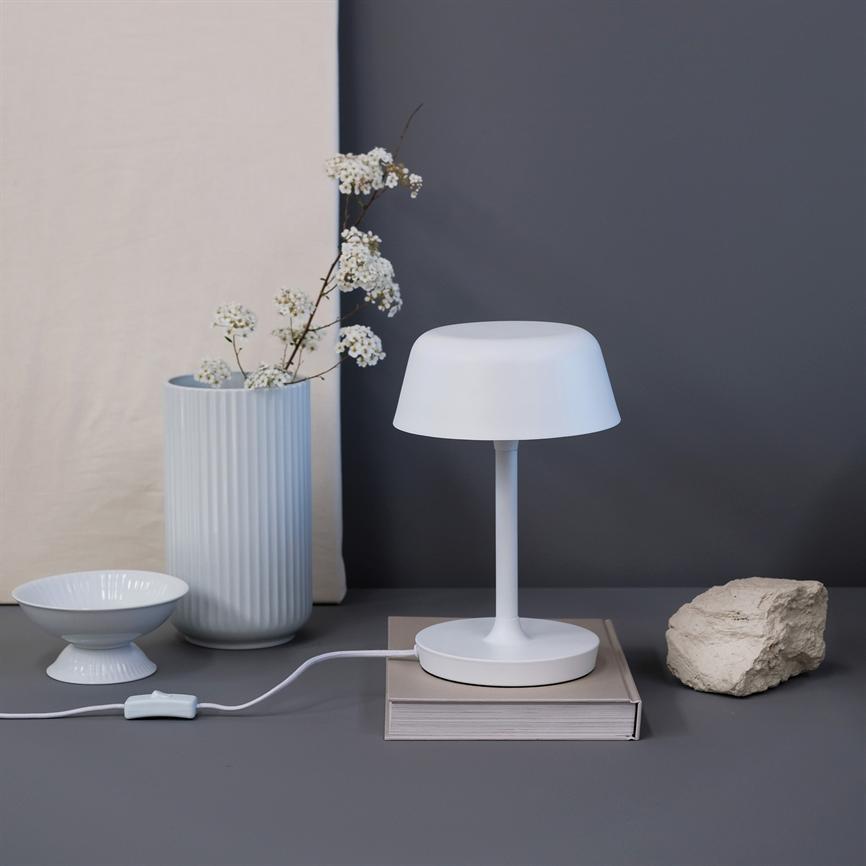 White table lamp stands on a book with visible cord, on a gray surface in front of a dark gray wall; on the left a fluted vase with white flowers and a bowl, on the right a rough stone.