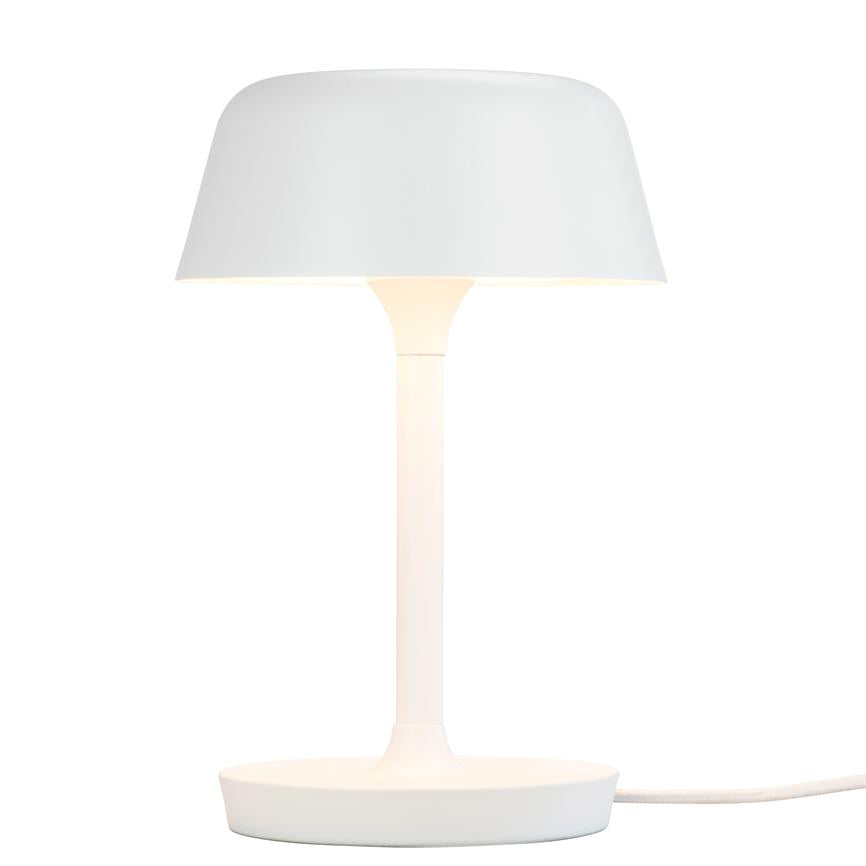 A white table lamp is lit; the screen casts soft light over a slim column and round foot, surrounded by a uniform white background.