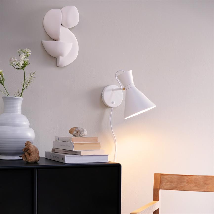 Wall lamp shines down on a black chest of drawers with stacked books, a white vase and small decorative stones; abstract white ceramic wall decoration hangs over a bright, minimalist space.