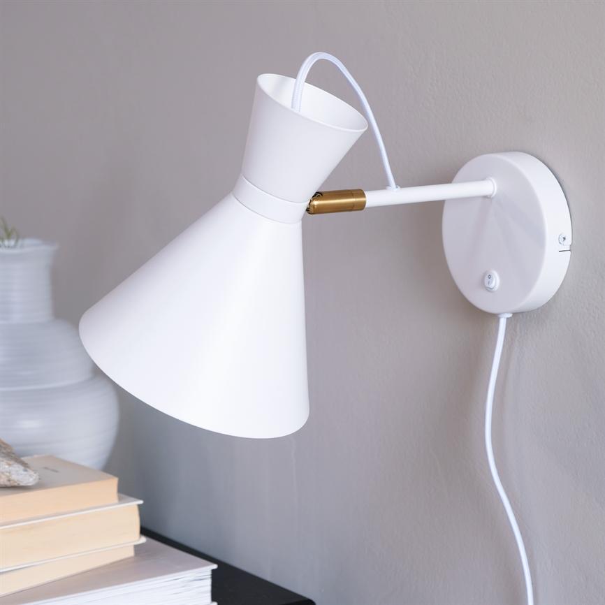 White wall lamp with conical shade is mounted on a gray wall and points down towards a stack of books on a black table; white wire runs down from the wall base.