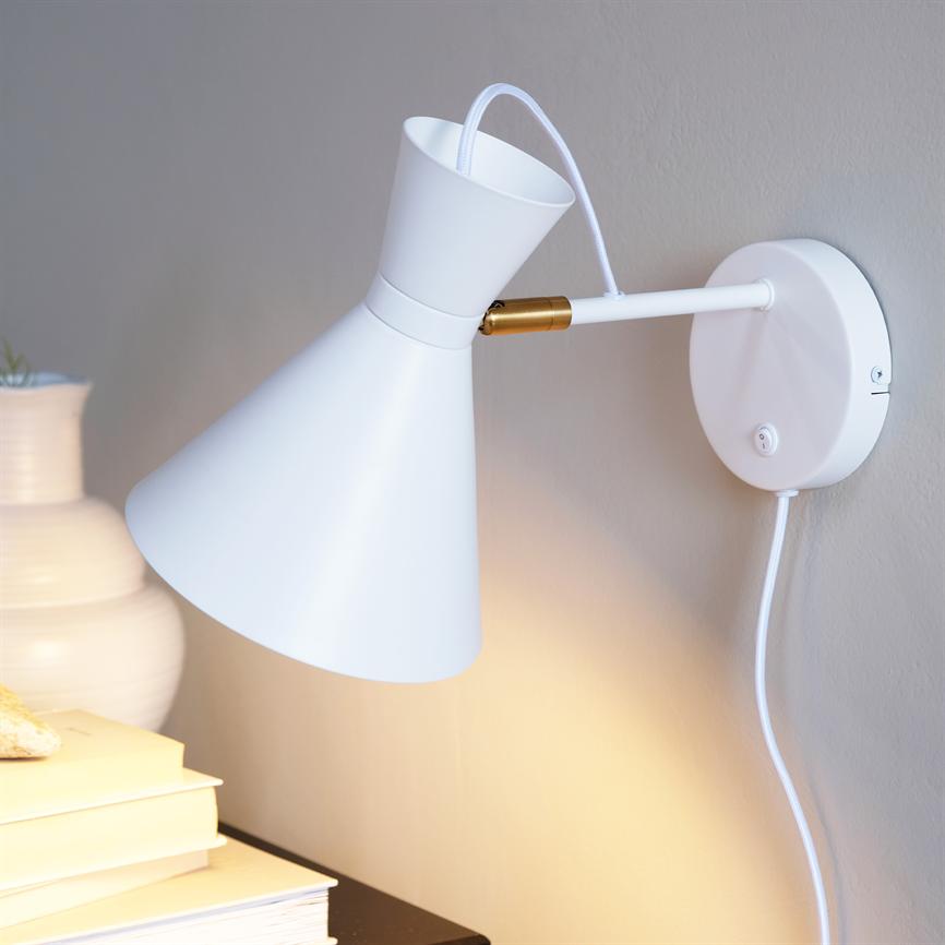 White metal wall lamp is mounted on a beige wall, angled downwards and casts warm light over a bedside table with a stack of books and a white vase.
