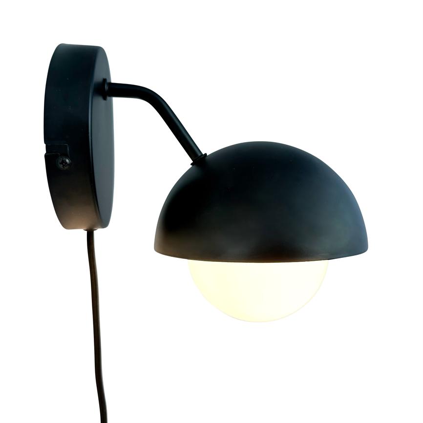 Black metal wall lamp with hemispherical shade, casting soft warm light from a white globe bulb; mounted on a round wall bracket with a visible black cable against a white background.