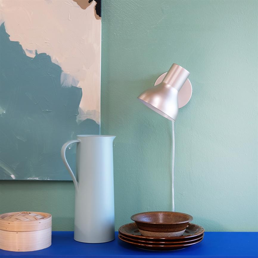 Wall lamp mounted on mint green wall, pointing down above blue shelf; on the shelf a tall pale blue jug, bamboo basket and stack of brown plates with a bowl; abstract painting on the left.