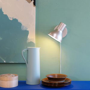 A white jug stands on a blue shelf; a wall lamp casts warm light over the jug and a stack of brown plates with a bowl in front of an abstract painting on a mint green wall.