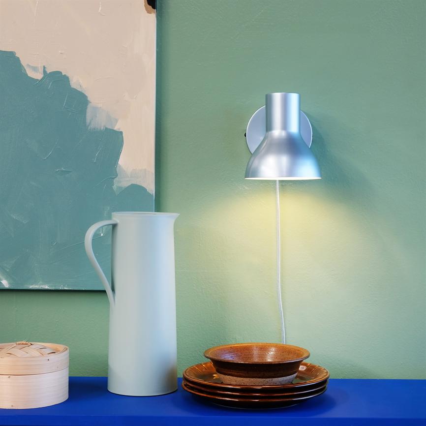 Wall lamp casts soft, warm light over a green wall; on a blue shelf is a tall pale blue jug, a small bamboo basket and a stack of brown ceramic plates.