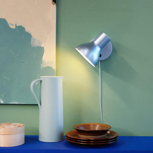 A silver wall lamp illuminates a green wall; on a blue shelf is a tall white jug, a small bamboo container and a stack of brown bowls and plates.