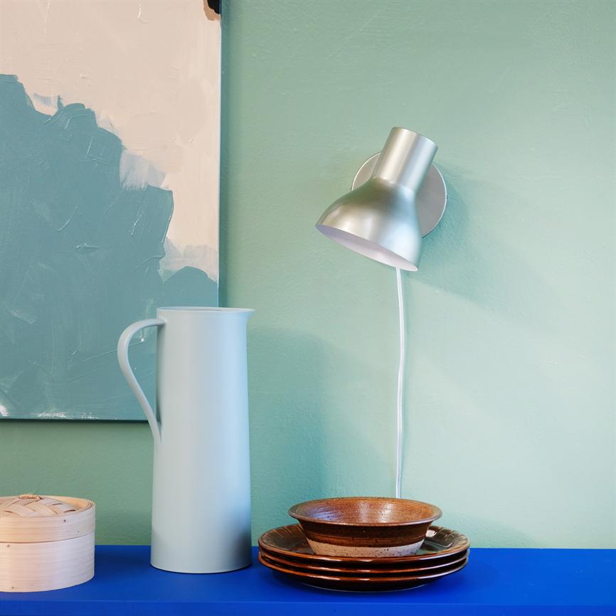 White jug stands on a blue shelf; a silver wall lamp shines down over the jug and a stack of brown plates and bowls, next to a bamboo steam basket, in front of a mint green wall.
