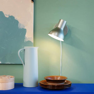 Wall lamp shines down on a white ceramic jug and stacked brown plates on a blue shelf, in front of a mint green wall with an abstract painting.