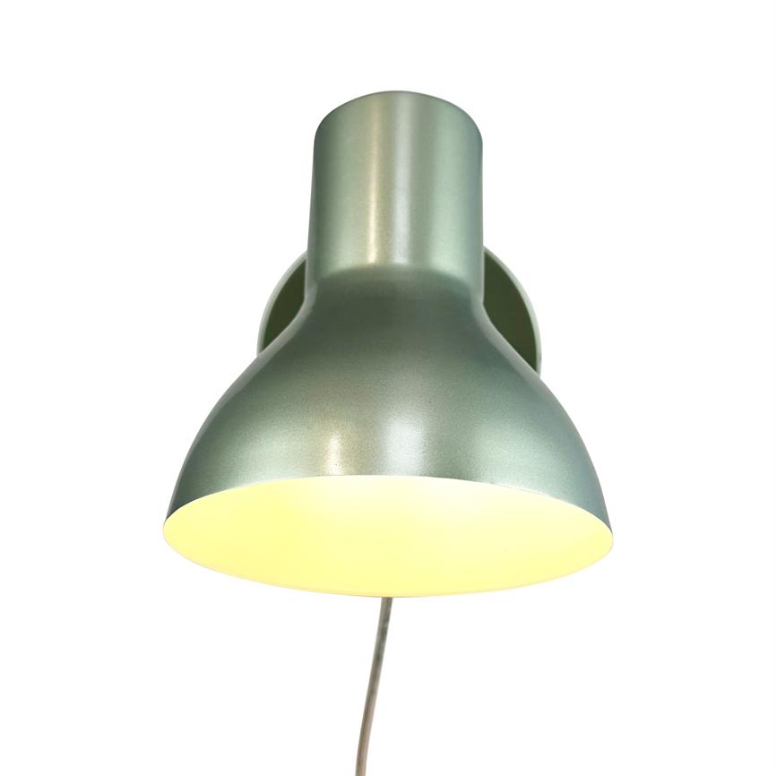 Green metal wall lamp shines downwards, shiny surface and warm yellow bulb glow; mounted on wall with round base, visible cord hangs down towards the floor, white background.
