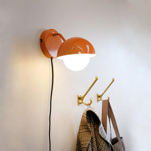 Orange wall lamp lights downwards, hangs from black cord; below it sit two brass hooks with a checkered coat and a brown bag on a light wall.
