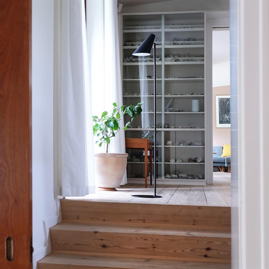 A floor lamp is tilted towards the floor by a large potted plant in a bright room with a wooden staircase, a glass display cabinet filled with small stones and a sofa in the background.