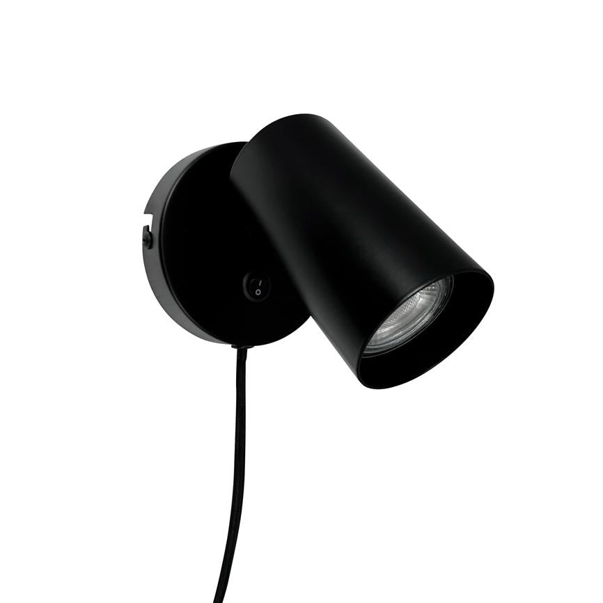 Black wall lamp with cylindrical shade, angled downwards, mounted on round wall base with visible cord and small switch, placed against a white, blank background.