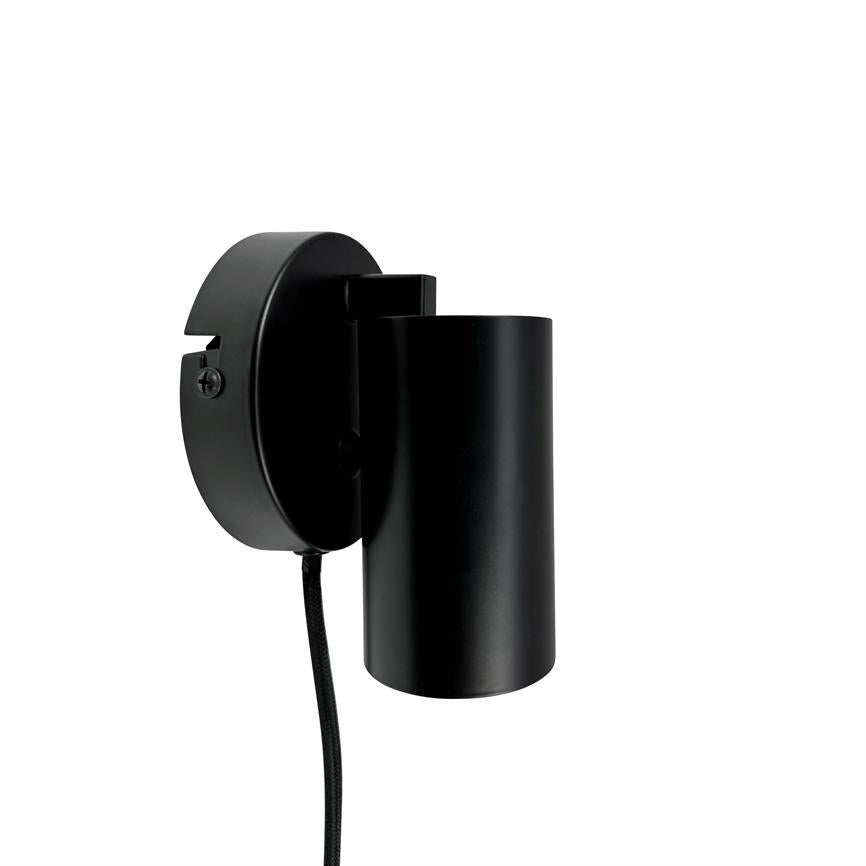 Black cylindrical wall lamp mounted on round wall bracket, with textile covered cord hanging down; minimalist design against a clean, white background.