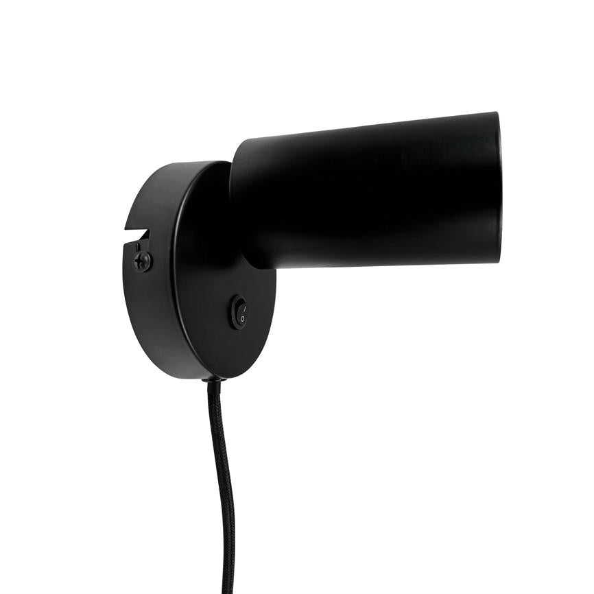 Black wall lamp points diagonally to the right, mounted on a round wall base with on/off switch; fabric-clad cord hangs down against a white background.