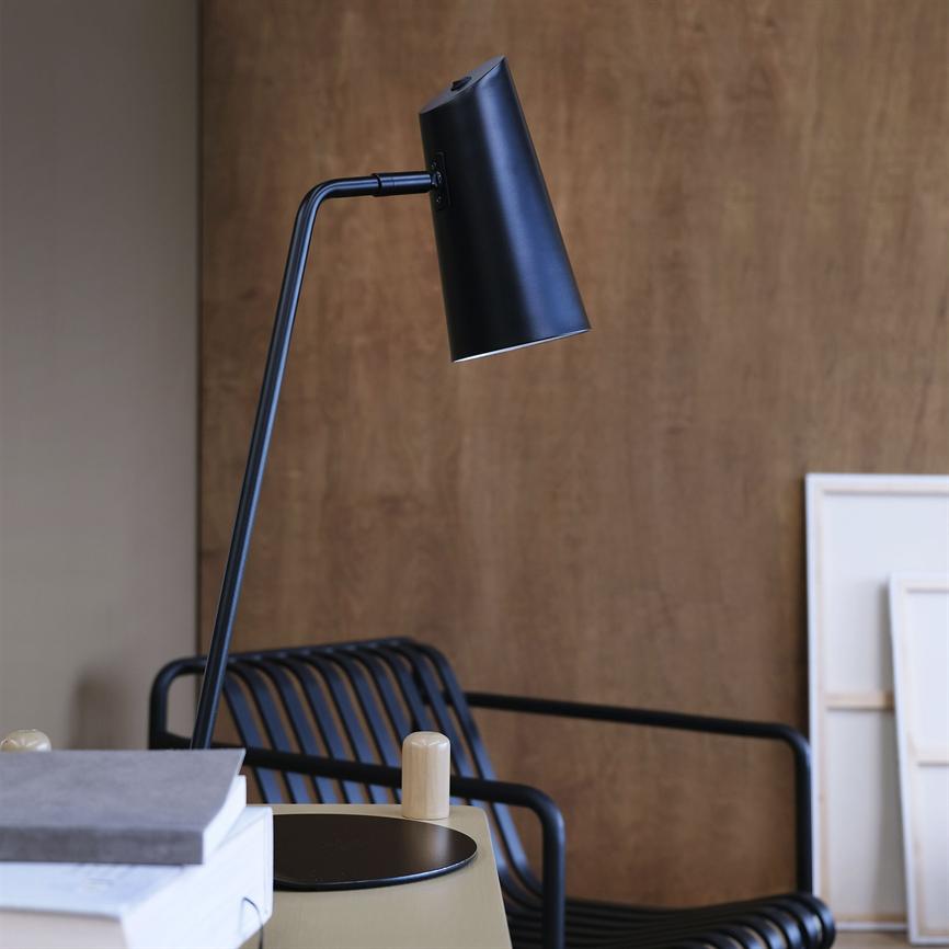 Black desk lamp points down above a side table in a modern interior; surrounded by stacked books, a black metal chair with slats and a wood paneled wall with upright canvases.