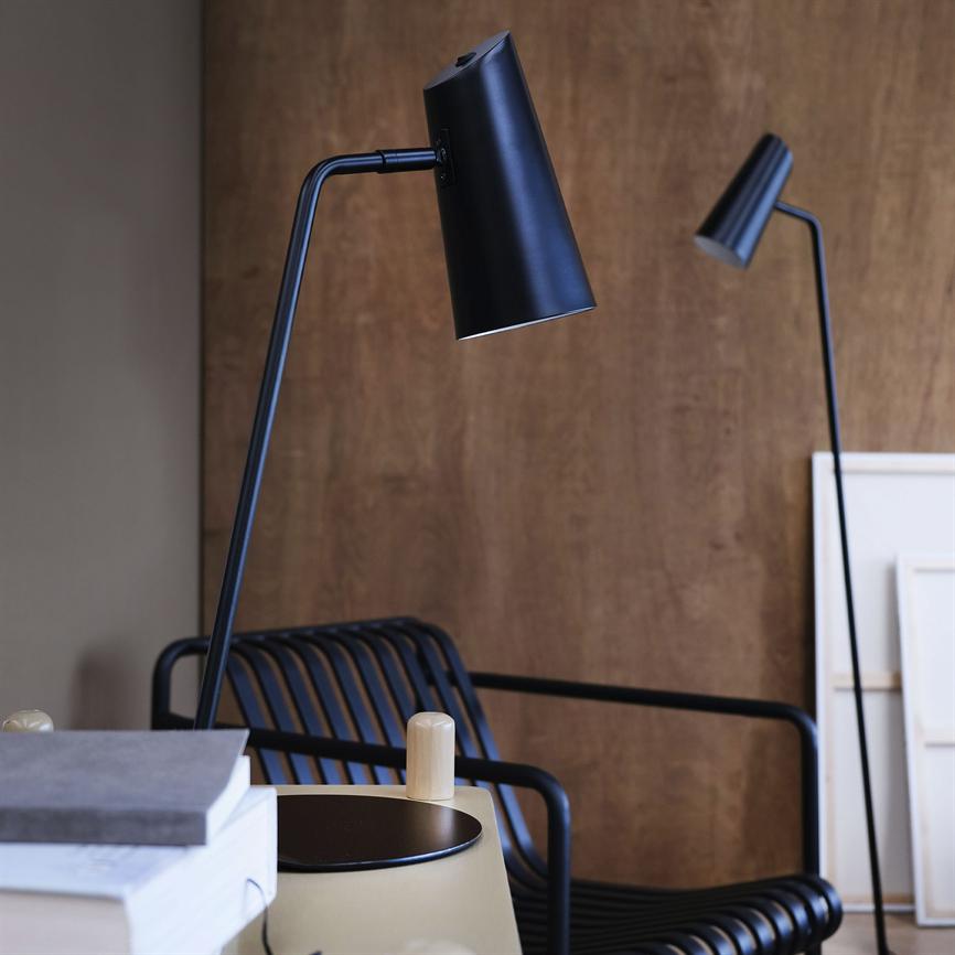Black reading lamp points down over a small table with stacks of books; a black ribbed metal chair and a slim floor lamp stand in front of a warm, wood-clad wall.