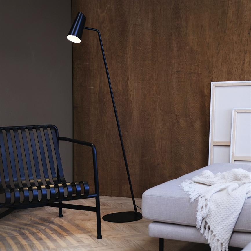 A black, slender floor lamp is lit and casts light on the floor; in a modern interior with a black metal bench on the left, a light gray sofa with a knitted rug on the right and a dark wooden wall.