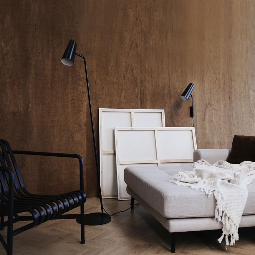 A light gray chaise longue with a knitted plaid draped over it stands in front of a dark wooden wall; two blank canvases lean against the wall, flanked by two black lamps and a black metal chair.