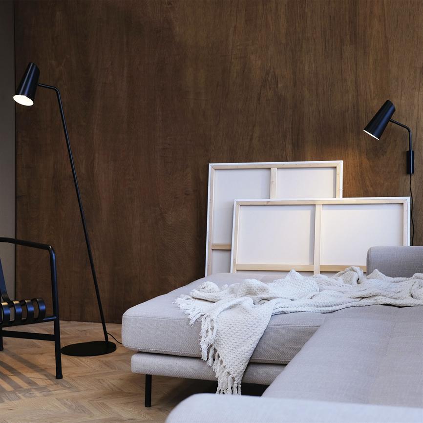 Low gray modular sofa has a white knitted rug; it stands in front of a dark wooden wall, two empty framed canvases lean against the wall and two black floor lamps illuminate the room.