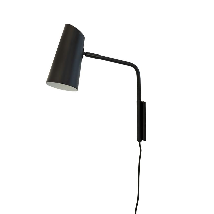 Black wall lamp with cylindrical shade, tilted forward; curved arm with rectangular wall bracket, power cable hanging downwards against a uniform white background.