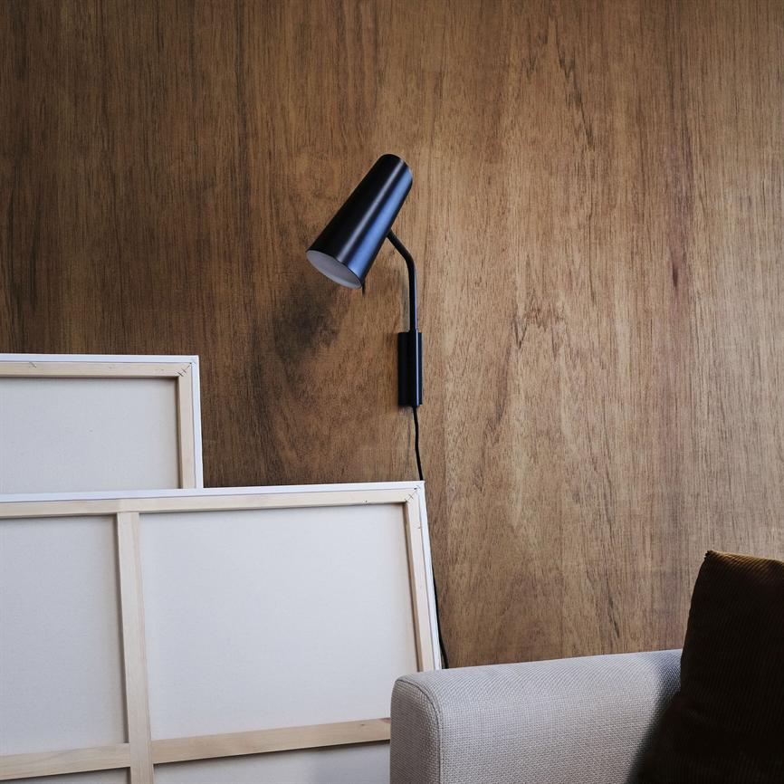 Black wall lamp mounted in a bent arm, facing downwards against a wood-clad wall; two blank canvases lean against the wall, with a beige sofa and dark cushion in the foreground.