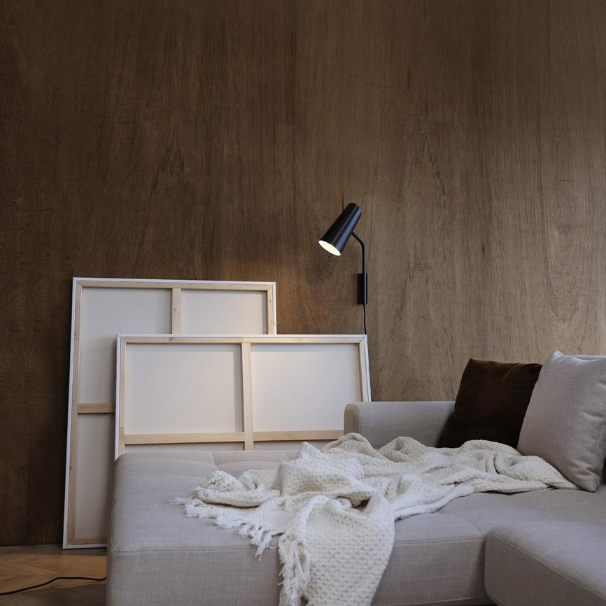 The sofa is covered with a crumpled, bright knitted rug; two standing canvases stand behind, while a lit black wall lamp casts warm light on a wood-clad wall.