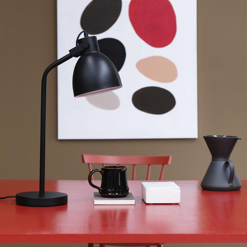 A black table lamp points down towards a red table top, where a black coffee cup on a base, a white box and a gray coffee maker stand in front of an abstract painting on a brown wall.