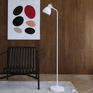 A white floor lamp stands upright next to a black lashing chair; in front of a dark wooden wall with an abstract canvas with oval color spots. Light gray carpet on a light wooden floor.