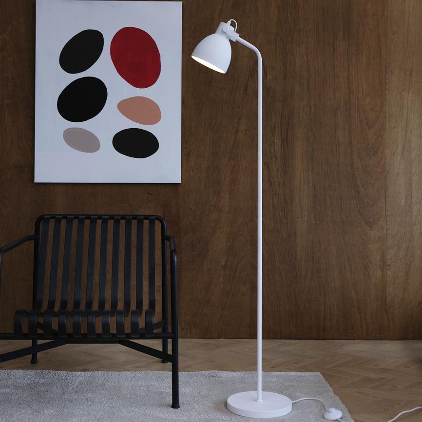 A white floor lamp is on and shines against a light carpet on the parquet floor, in front of a dark wooden wall with an abstract painting with colored ovals and a black chair with slats.