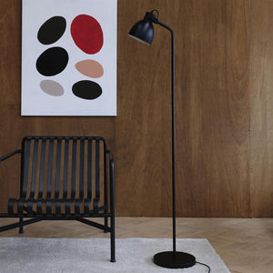 Black floor lamp stands upright and points obliquely downwards next to a black metal chair under an abstract painting on a wood paneled wall above a light wood floor and light carpet.
