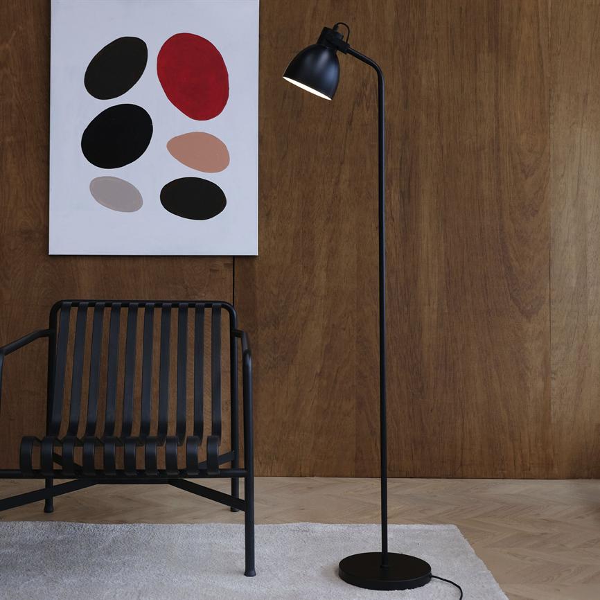 A black floor lamp is lit next to a black metal chair; behind it hangs an abstract painting on dark wooden panels, above a light carpet on a parquet floor.