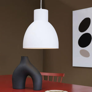 White hanging lamps hover above a red table, illuminating a black, curved vase; in the background brown wall with abstract painting and a red chair.