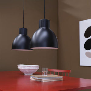 Two black domed hanging lamps hang above a red table with a stack of books and a glass; the room has brown walls, a red chair and a white bowl.