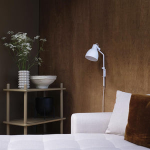 White wall lamp is mounted on a warm wooden wall above a light gray sofa; a small table to the side holds a beaded vase, white bowl and black pot with white flowers.