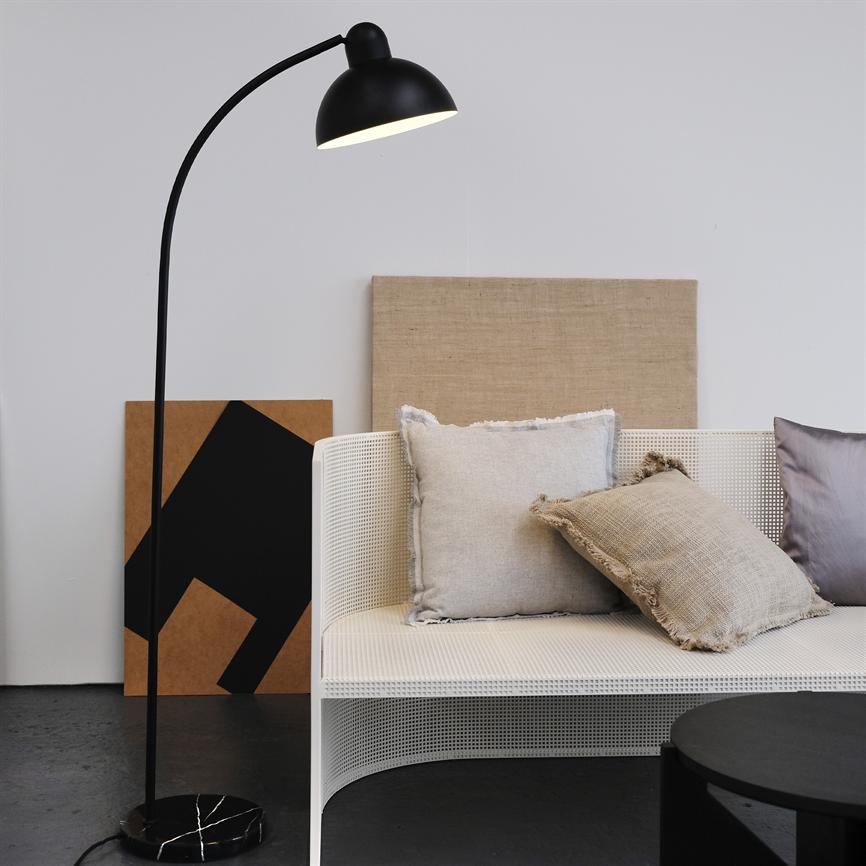Black arc-shaped floor lamp casts soft light over a white mesh sofa with three decorative pillows; behind it, two canvases are leaning against a light wall in a minimalist, quiet room.