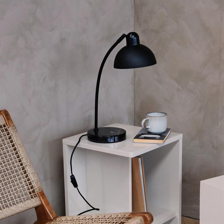 A black table lamp bends over a white bedside table; on the table is a ceramic cup and a bound book. Next to it, a wicker wooden chair stands in front of a gray textured wall.
