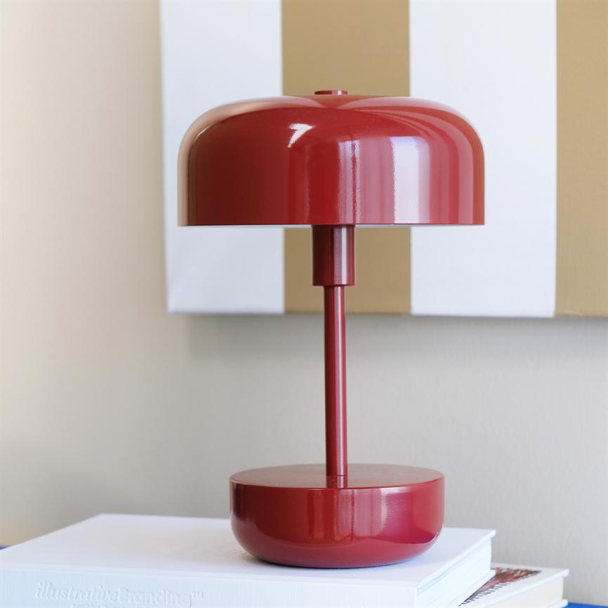 Red metal table lamp stands on a stack of books; glossy screen and slim base; the background is a wall with white and beige vertical stripes. Visible text on spine: illustrativeBranding™