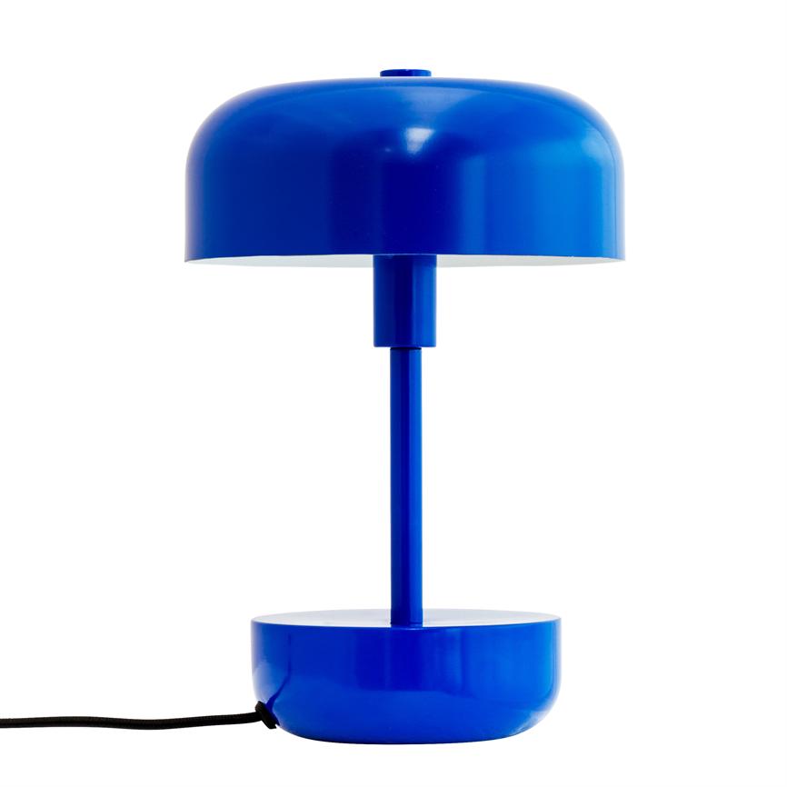 A blue metal table lamp with a round shade and cylindrical base stands in the middle of a white background; a black wire protrudes from the base of the lamp.