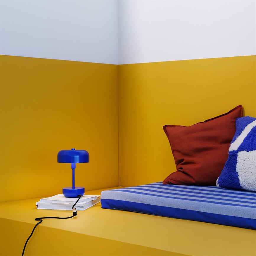 A blue table lamp stands on a stack of books by a yellow wall corner; on the right is a blue-striped mattress with a red pillow and a blue-white plush pillow.