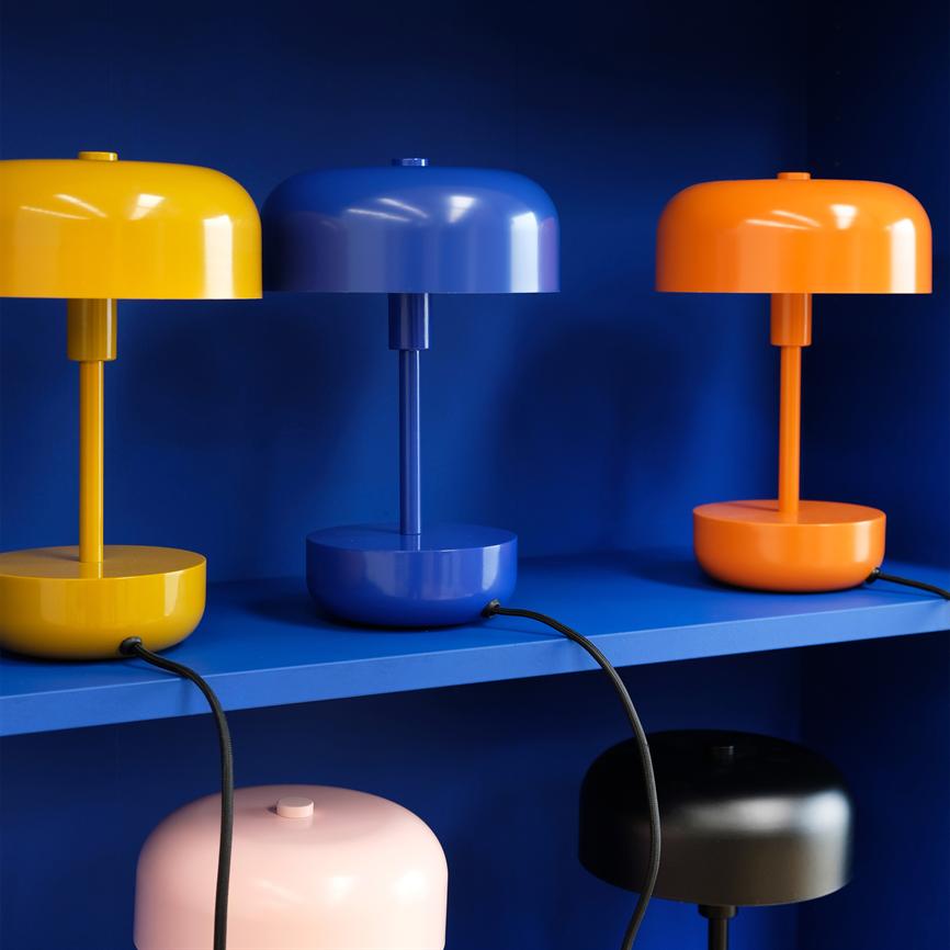 Three dome-shaped lamps (yellow, blue, orange) stand on a blue shelf; black cables hang down, back wall and shelf are dark blue, partially visible pink and black lamps underneath.