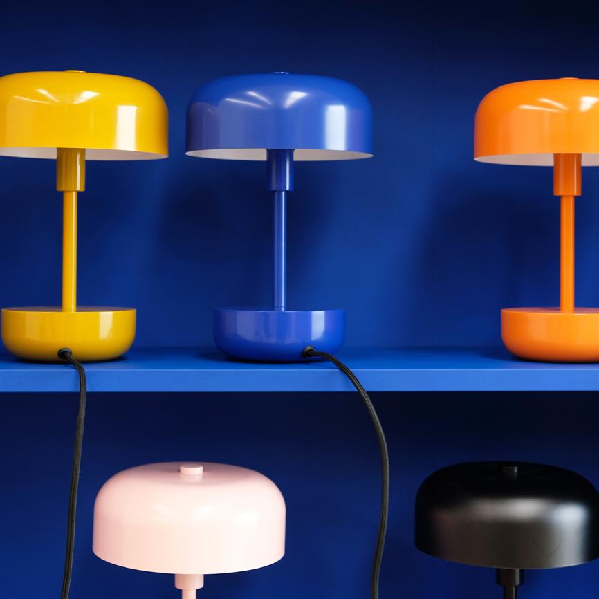 Multicolored table lamps (yellow, blue, orange at the top; pink, black at the bottom) stand on blue shelves; their black wires hang down in front of a deep blue background.