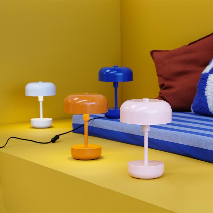 Four small, modern table lamps in white, orange, blue and pink stand on a yellow shelf in front of a blue-striped mattress and a brown pillow.