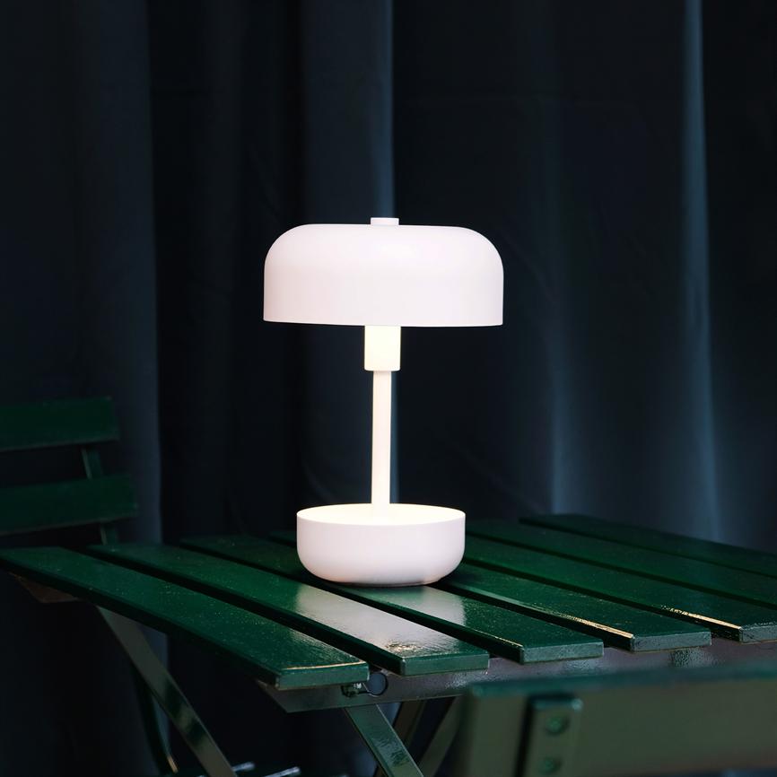 White table lamp stands and emits soft, warm light on a dark green folding wooden table; the background is dark with thick curtains.