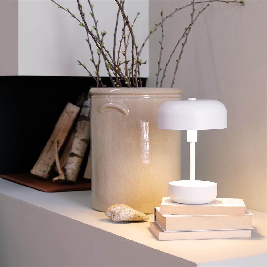 A white table lamp shines softly on a small stack of books; it stands on a shelf next to a large beige jar with slender spring branches and birch trunks.