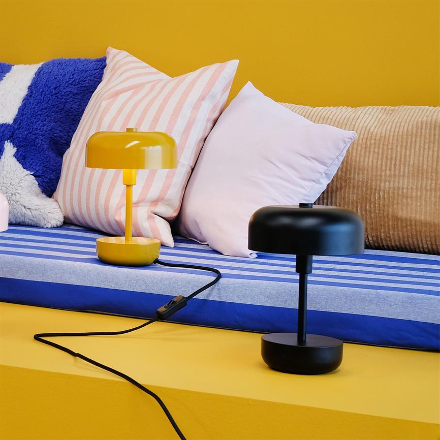 Two small lamps, one yellow and one black, stand on a blue-striped upholstered seat; soft cushions in pink, pink-striped, blue-white and beige lean against a yellow wall.