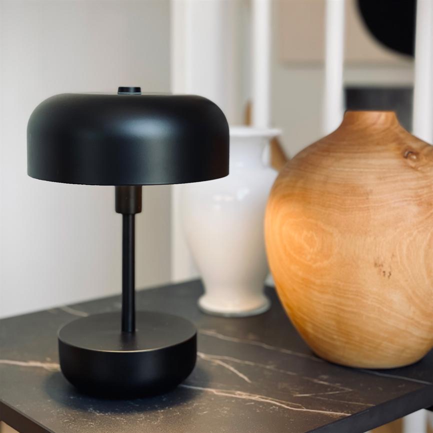 A black table lamp stands on a dark marble table, flanked by a white vase and a round wooden vase in a bright, modern corner of the living room.