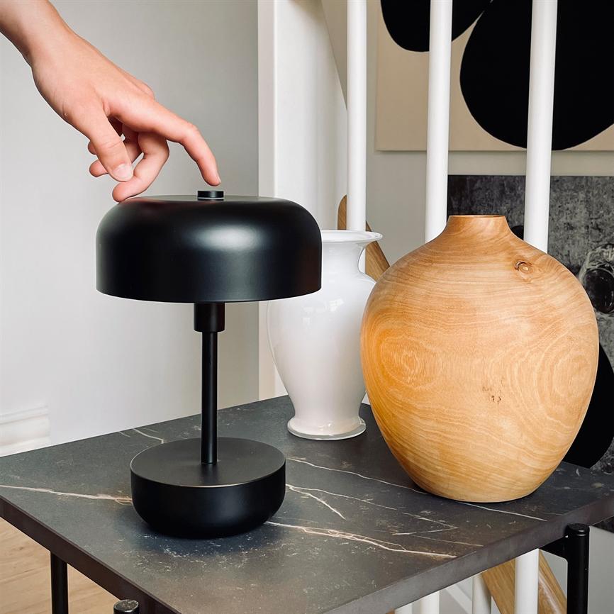 Black table lamp, a hand reaches out and presses its top; stands on a dark marble table between a white vase and a round wooden vase in front of white plays and wall art.